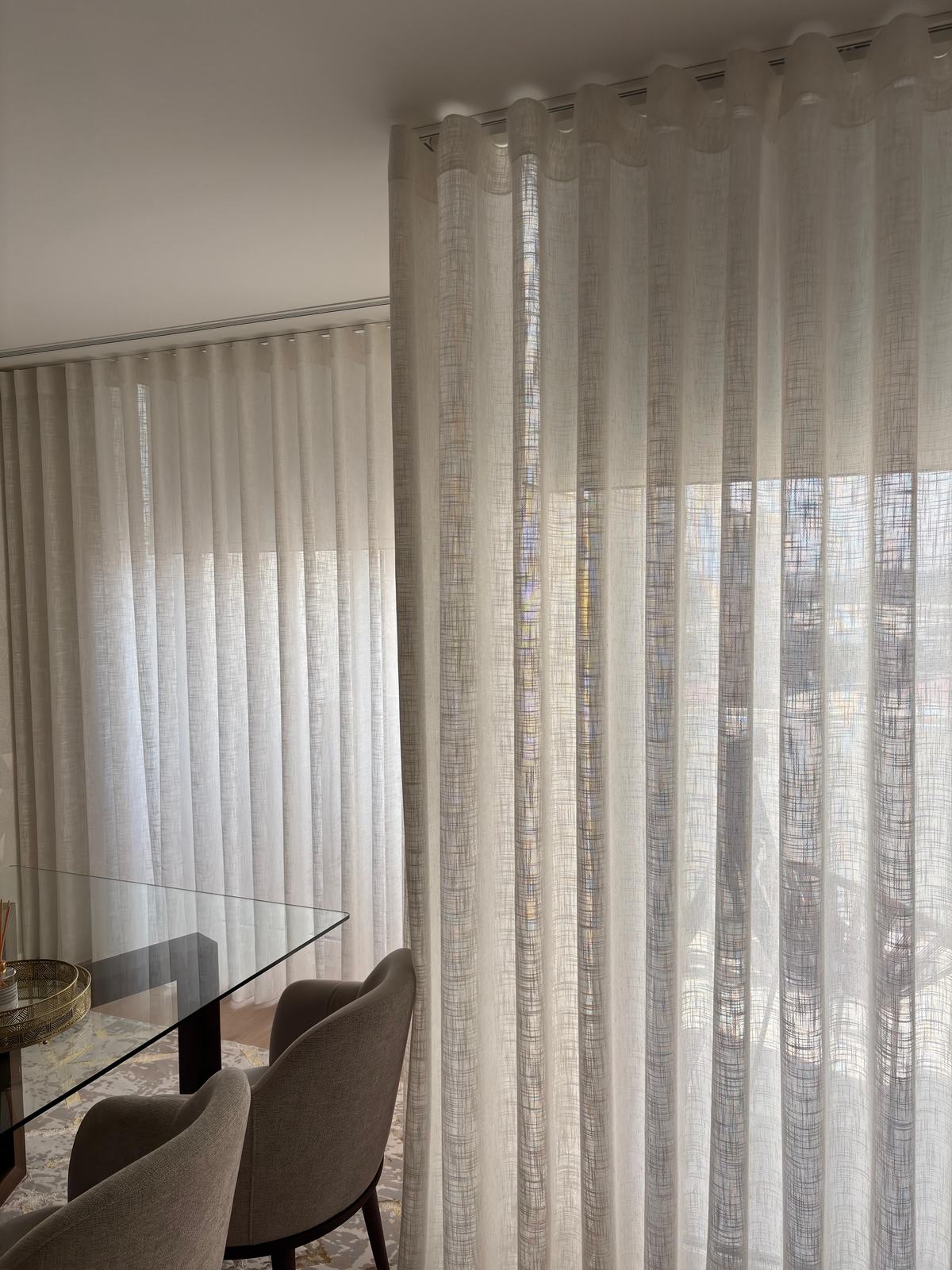 Cortinado de onda em linho numa sala de jantar, visto através da mesa de vidro