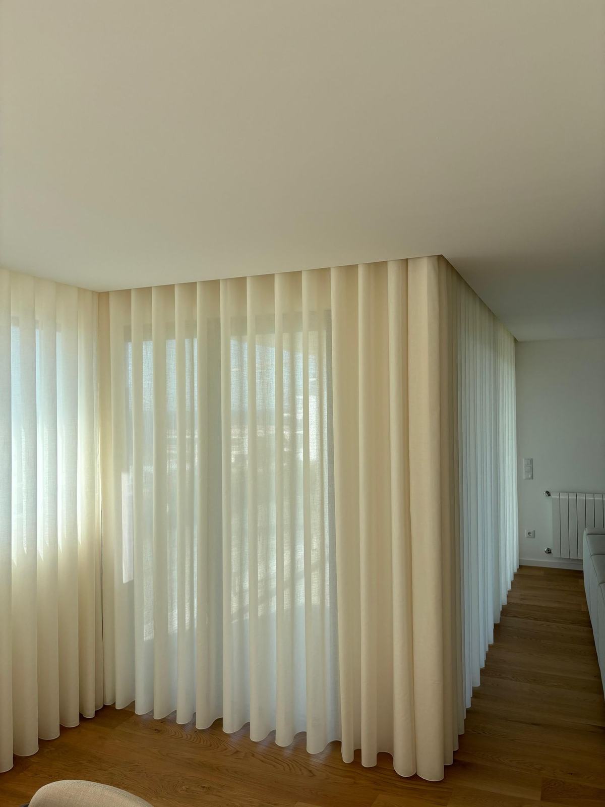 Cortinado de onda em tom creme a acompanhar um corredor com chão de madeira