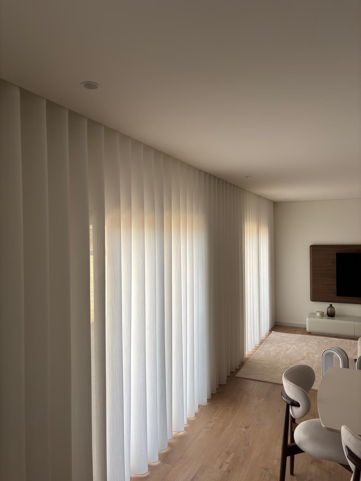 Cortinados de onda em tecido claro a percorrer uma sala aberta com chão de madeira