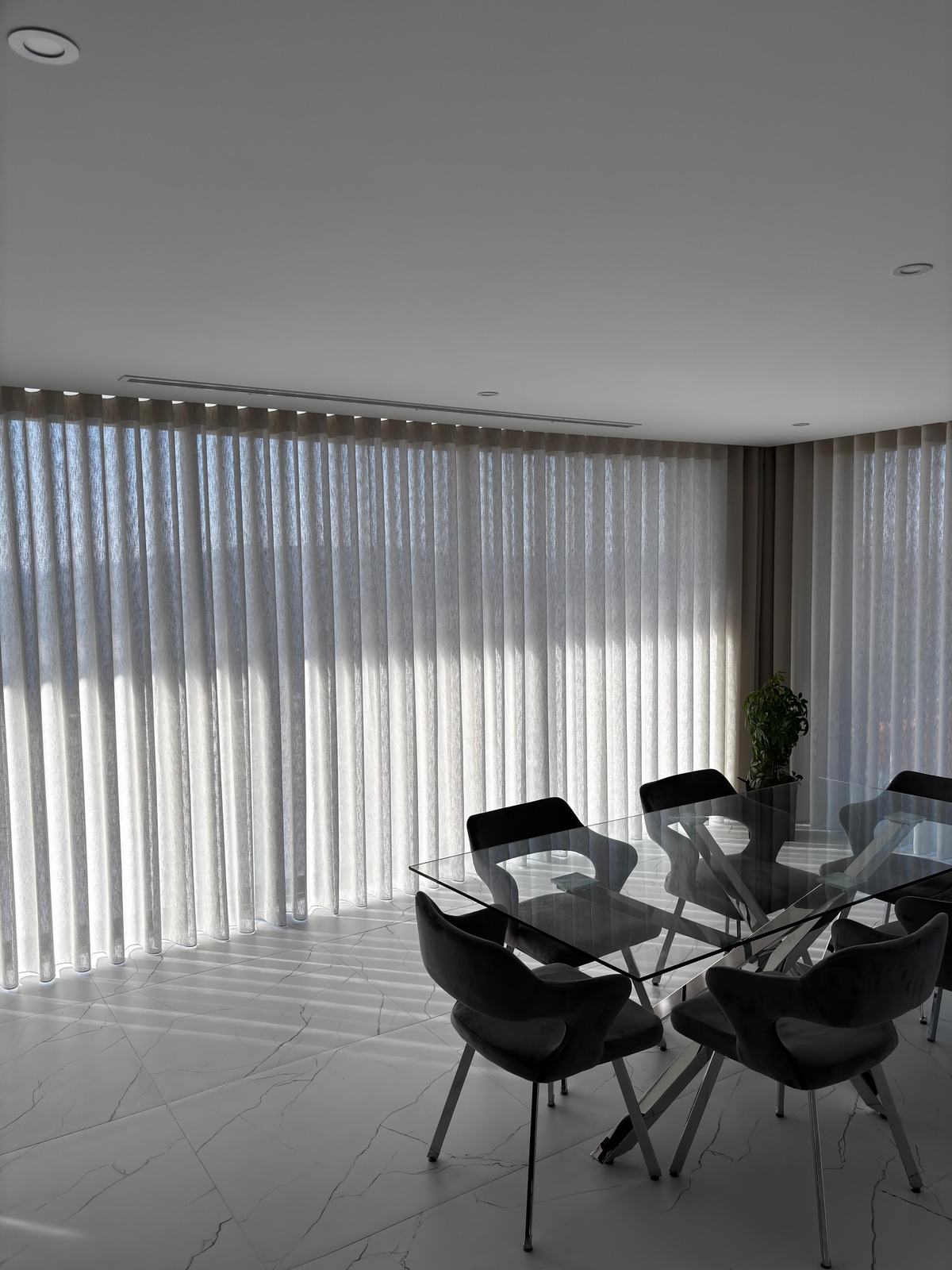 Sala de jantar com mesa de vidro e cadeiras pretas diante de cortinados de onda